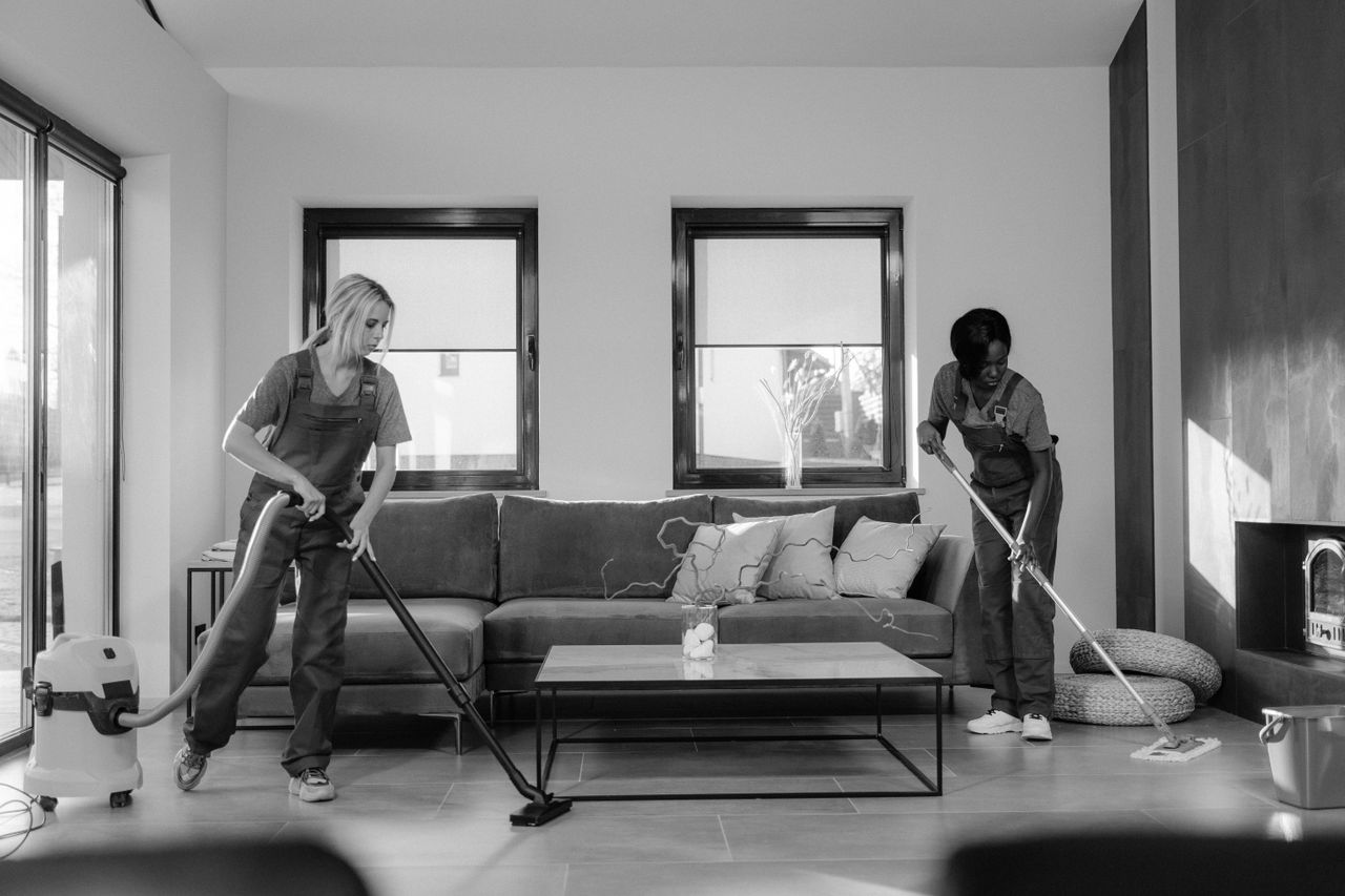 services-03 Two women in work clothes tidying a modern living room with a vacuum and a mop.